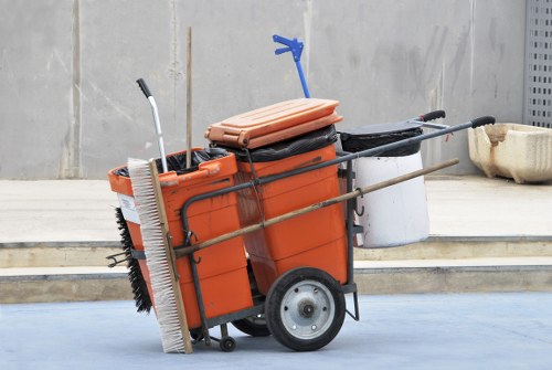 A mobile commercial waste collection cart positioned on a paved surface outdoors, with a concrete or stone wall in the background. The cart features a large orange plastic container with a black lid, mounted on a metal frame with two large rubber wheels at the rear and small support legs at the front. Attached to the cart are various cleaning tools, including a broom with bristles facing downward, a mop with a white handle, and a small dustpan, all arranged in a practical manner. A folded orange plastic bag or tarp is placed on top of the container, and a blue clip or clamp is attached to it. To the right, a black hose or pipe extends from the cart toward a white bucket or container, resting on the ground. The environment suggests an outdoor area associated with commercial waste collection or rubbish removal, consistent with services offered by Commercial Waste Northolt, near postcode NW10, in London.