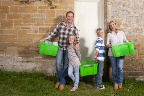 A group of four people, including two adults and two children, stand outdoors against a rustic stone wall, each holding bright green plastic storage bins used for rubbish collection or disposal. The man on the left, dressed in a checkered shirt and jeans, smiles and holds a bin with his right hand while a young girl, wearing a patterned sweater and jeans, stands close to him, also holding a bin and smiling. To the right, a boy in a striped shirt and jeans stands next to a woman in a light-colored top and jeans, who is holding a green bin at waist level. The setting appears to be a residential or semi-urban area, with a grassy patch in the foreground and a white door set into the stone wall behind them. The scene suggests an organized rubbish removal or waste management activity, aligning with services offered by Commercial Waste Northolt, a company specializing in rubbish and business waste removal in the Northolt area. The natural lighting and casual composition emphasize a straightforward, community-oriented approach to waste disposal solutions.