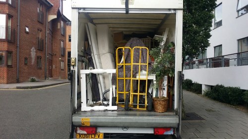 The image shows the interior of a small commercial waste collection vehicle parked on a city street, with the rear doors open. Inside, there are various discarded items including a yellow hand truck leaning against a stack of furniture and miscellaneous waste materials. Among the objects visible are a white plastic pipe, a small woven basket filled with greenery or plant material, and different pieces of furniture or equipment, possibly including parts of office or household items. The vehicle's interior is lined with metal and appears to be designed to hold waste or bulky refuse. The setting includes a paved sidewalk, modern residential buildings with brick and white facades, and some greenery. The scene suggests a professional rubbish removal service operating in an urban area, likely related to commercial or residential waste collection in Northolt or a nearby locality.