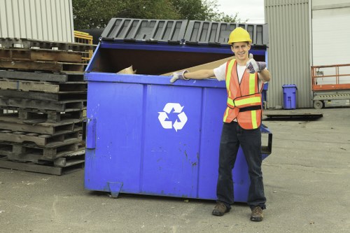 A male waste management worker wearing a yellow safety helmet, high-visibility orange and yellow vest, and gloves, standing in an industrial area near a large, blue metal skip with a partially open black lid. The skip features a white recycling symbol on its front and is situated on a paved surface, with stacks of wooden pallets to the left and a metal shed or warehouse in the background. The worker is smiling and giving a thumbs-up gesture, indicating successful rubbish collection or disposal. The setting suggests a commercial or industrial waste removal operation, possibly servicing businesses in Northolt or nearby areas. The environment is well-lit with natural daylight, and the area appears organized, with additional waste containers and equipment visible in the vicinity, supporting the context of professional rubbish removal services.
