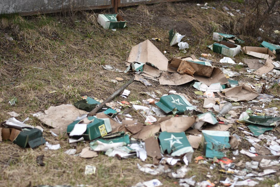An outdoor scene shows a patch of grass and soil near a slope, scattered with various debris and waste. There are multiple torn and crumpled cardboard boxes, predominantly green with white star logos,