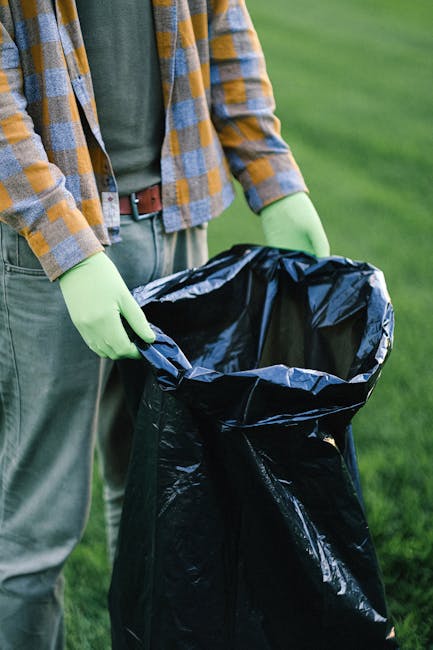 An outdoor scene shows a patch of grass and soil near a slope, scattered with various debris and waste. There are multiple torn and crumpled cardboard boxes, predominantly green with white star logos,